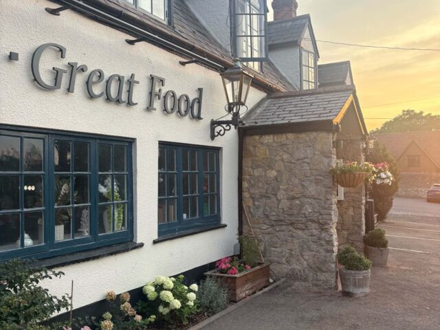 The front of The Horseshoe at sunset: white walls, dark window frames, hanging baskets and Great Food on the wall