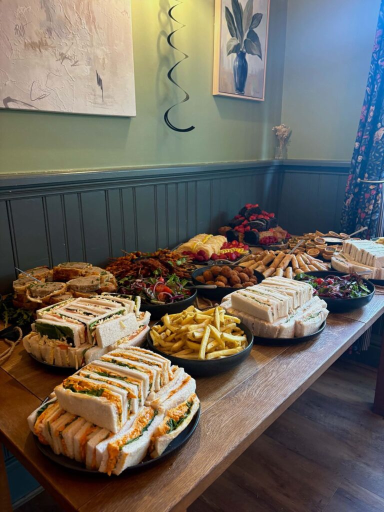 A party buffet along a table in the dining room: plates of sandwiches, bowls of chips and salad, sausage rolls, tarts and strawberries